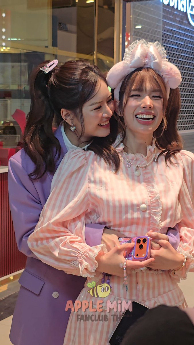 Two smiling women pose together indoors, one wearing a pink and white striped dress with a decorative bow, and a watermark reading "APPLE MIM FANCLUB TH" appears at the bottom.