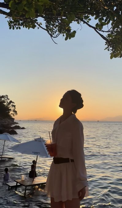 A silhouetted woman stands on a beach at sunset, holding a drink and looking out over the water, with beach umbrellas and a tree branch framing the scene.