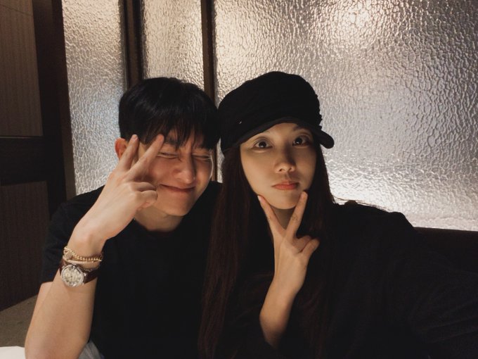 A selfie of a young couple both making peace/V signs; the man on the left wears a black t-shirt and has his eyes closed with a smile, while the woman on the right wears a black cap and black top, sitting indoors near a frosted glass window.