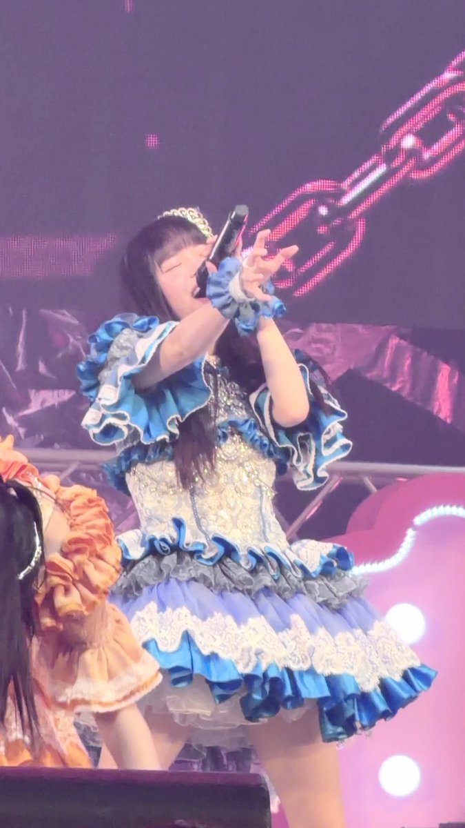 A young woman performing on stage in an elaborate blue and white frilly dress with a small crown, holding a microphone up to her face with her eyes closed.