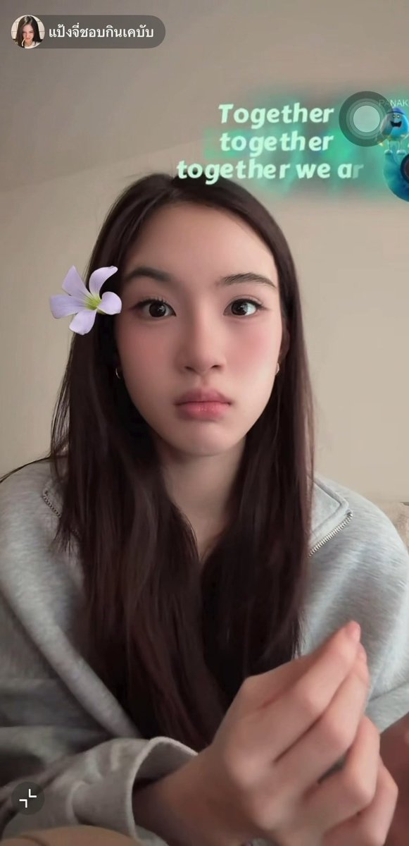 A selfie of a young woman with long dark hair wearing a gray top, featuring a digital purple flower filter on her cheek, green karaoke-style text overlay ('Together toggether together we ar...'), and Thai text 'แป้งงี้ขอบกินเคบับ' at the top.