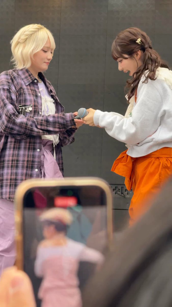 Two young women, one in a purple plaid shirt and the other in a white hoodie, exchanging a microphone on a stage while a phone records them in the foreground.