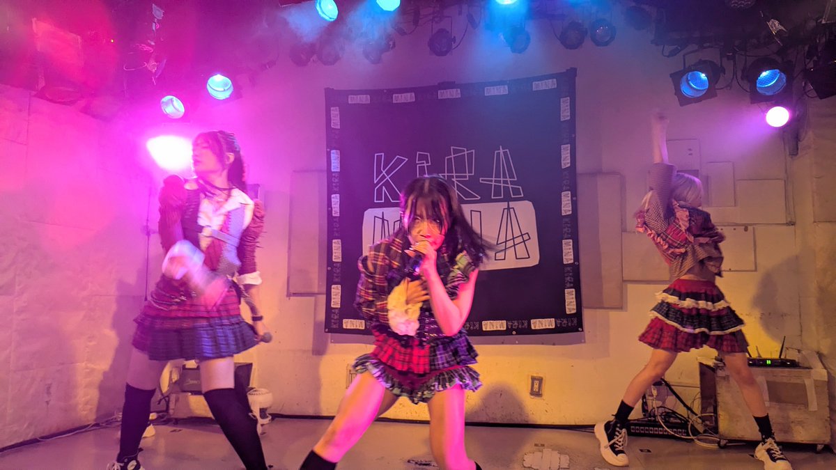 Three performers in plaid skirts and dark tops perform on a stage illuminated by purple and pink lights, in front of a banner that reads 'KiKA KiKA'.