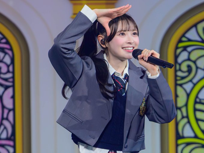 A young female performer on stage wearing a grey school uniform blazer, holding a microphone and posing with her hand near her head.