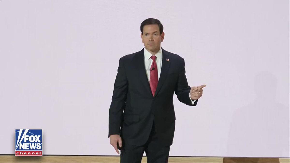 A man in a dark suit with a red tie and an American flag pin gestures with his hand while standing in front of a white background, with a 'FOX NEWS channel' logo in the bottom left corner.