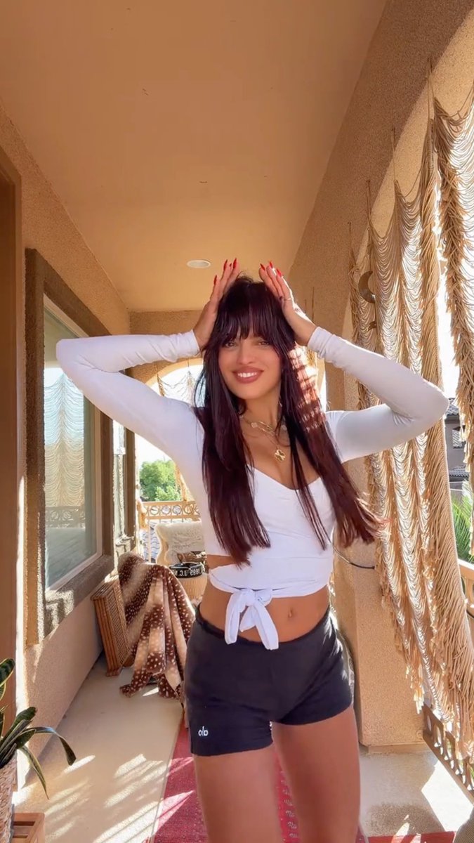 A smiling young woman with long dark hair and bangs poses with her hands on her head, wearing a white long-sleeve tied crop top, black Alo shorts, and gold jewelry on a sunlit balcony.