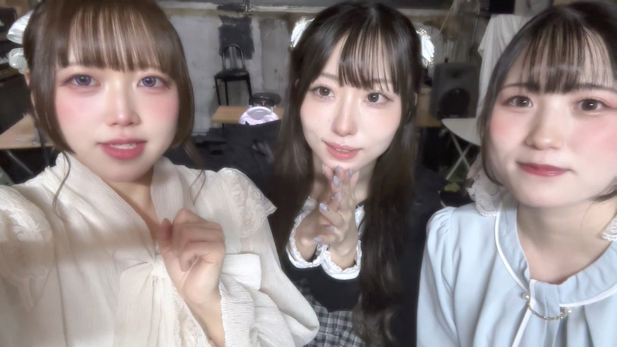 Three young women pose closely together for a selfie, with the two on the ends showing stylized makeup and the middle one clasping her hands together.