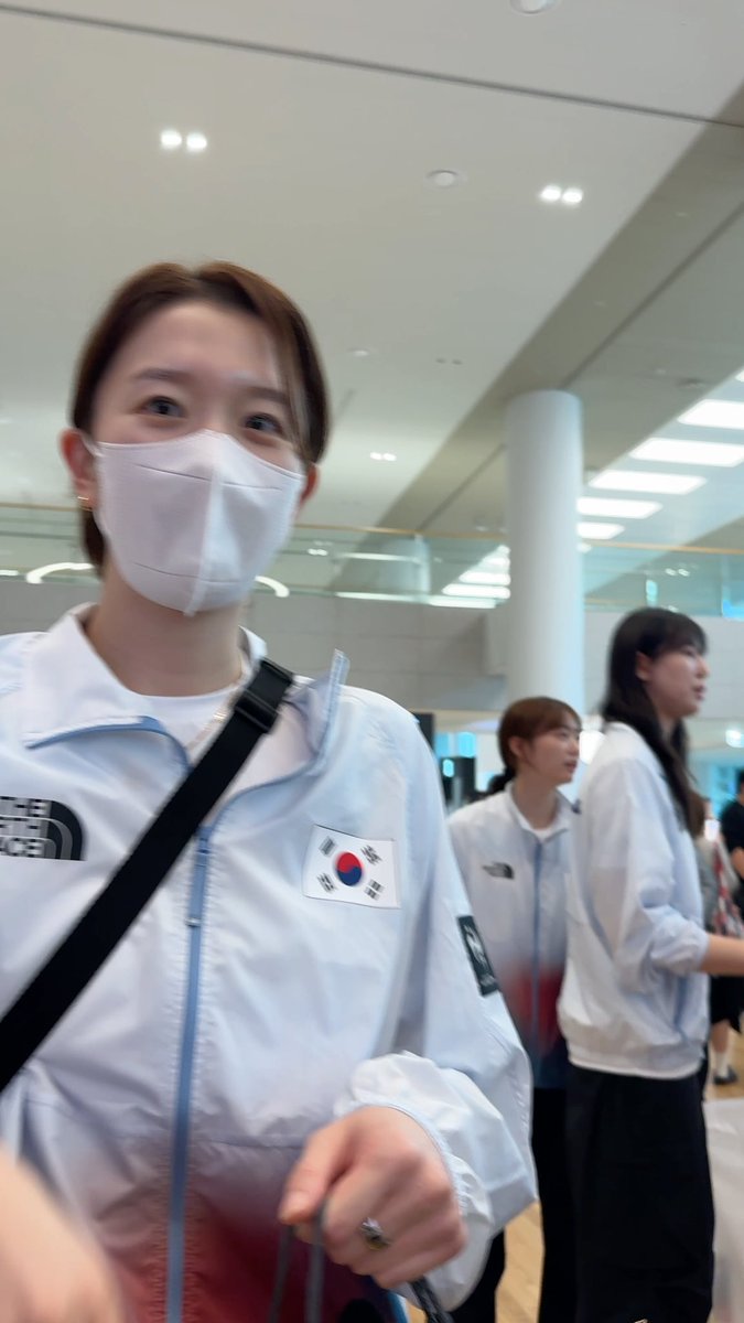 A young woman wearing a white face mask and a light blue North Face jacket with a South Korean flag patch on the chest is seen in what appears to be an airport or transit terminal, with other people in similar team jackets in the background.
