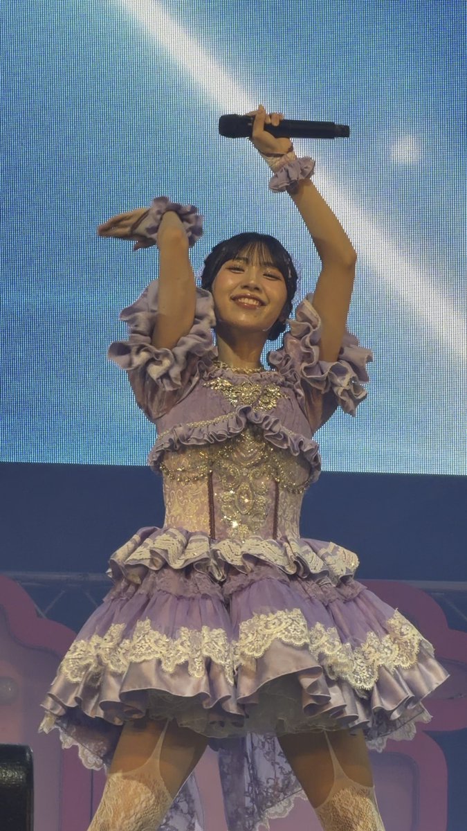 A young woman performer on stage in an elaborate lavender ruffled dress with a corset, holding a microphone and smiling brightly against a large blue screen.