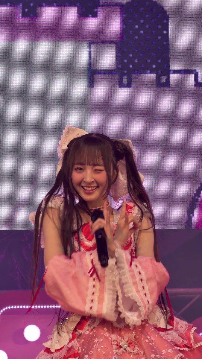 A young woman performer on stage in a pink ruffled idol costume with pigtails, holding a microphone and winking at the camera.