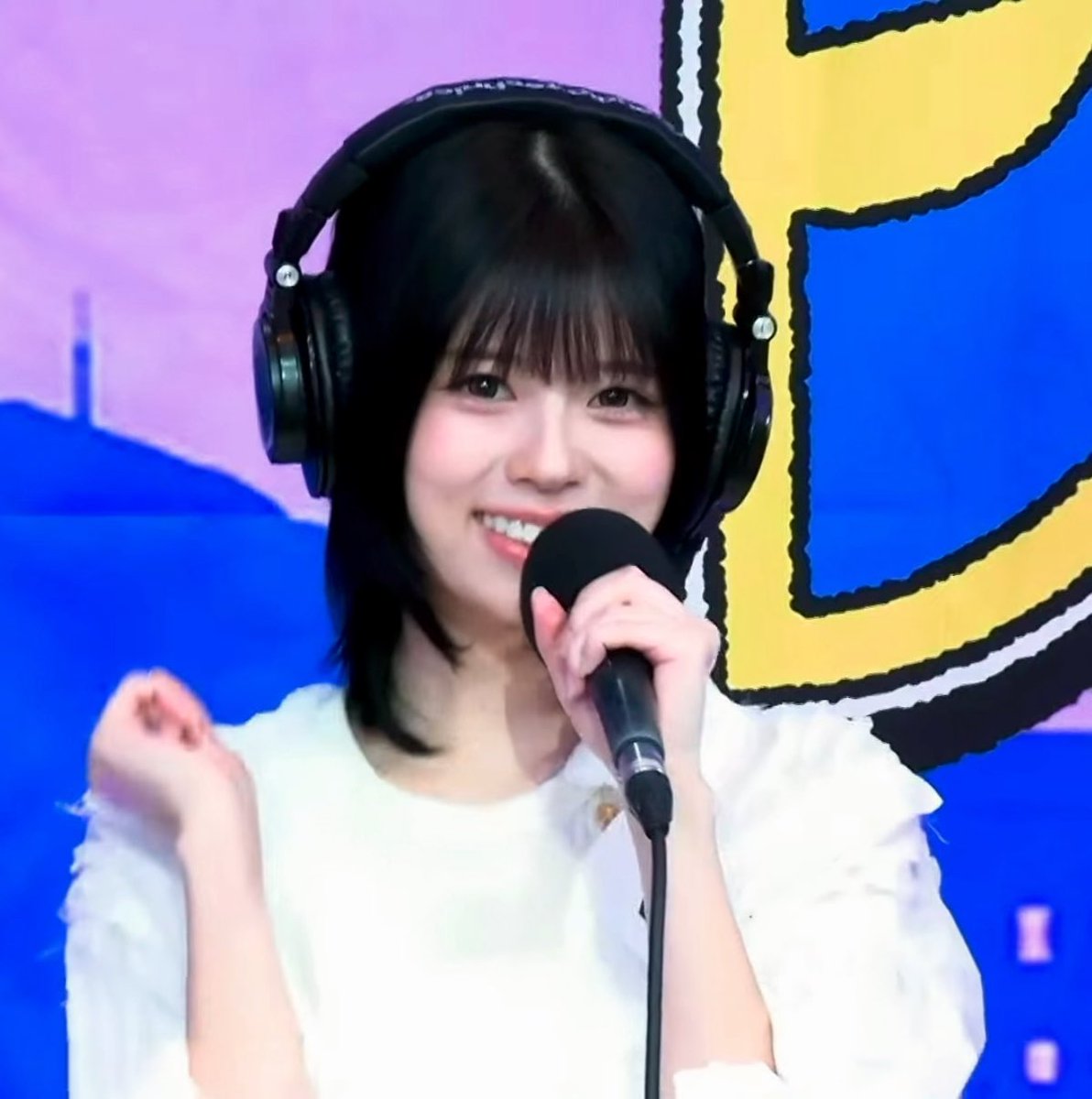 A young woman with dark hair wearing large headphones and a white top, smiling into a microphone against a blue and yellow background.
