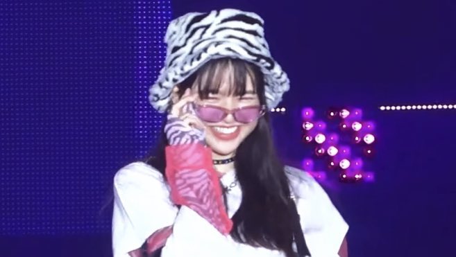 A K-pop performer wearing a zebra-print bucket hat, pink sunglasses, and a white outfit, smiling on a stage with purple lighting.