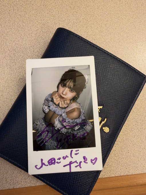 A polaroid photo of a young woman in a frilly dress, signed with purple marker and Japanese text, resting on a dark blue leather case.