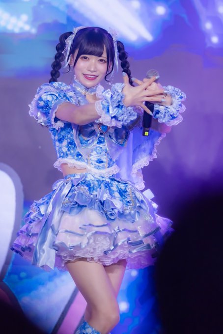 A young woman performs on stage wearing an elaborate blue and white frilly idol costume, with her hair styled in twin braids, holding a microphone as she sings.