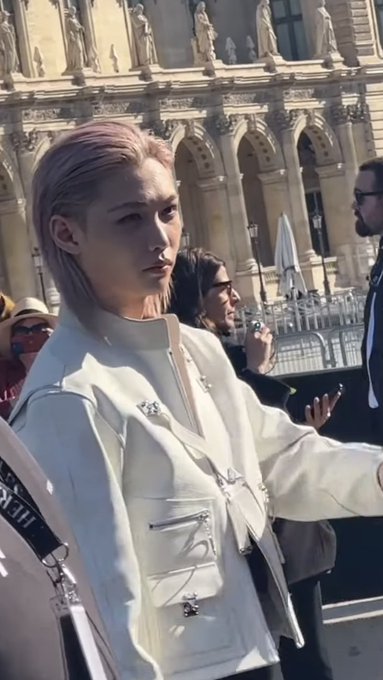 A stylish individual with short pinkish hair wearing a white jacket with silver hardware poses outdoors in front of an ornate classical building.