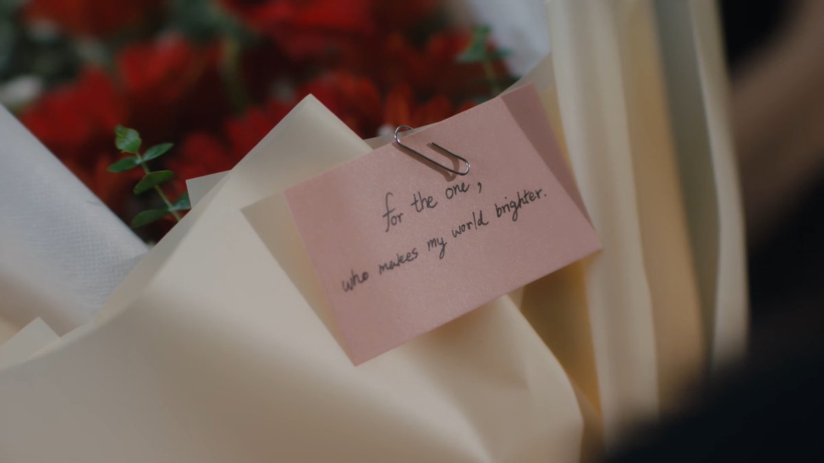 Close-up of a bouquet of red roses wrapped in cream-colored paper, featuring a pink note attached with a paperclip that reads 'for the one, who makes my world brighter.'