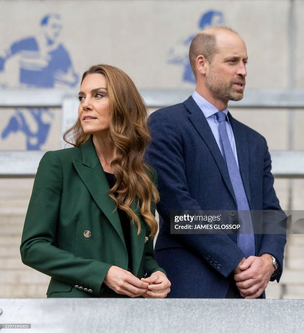 The Prince and Princess of Wales stand together at AMT Headingley Stadium during their visit to the home of the Leeds Rhinos rugby team. The woman wears a green blazer while the man is dressed in a navy suit with a blue tie, as they observe activities at the venue where they learned about the Leeds Rhinos Foundation's work using sport to change lives. The image is credited to James Glossop.