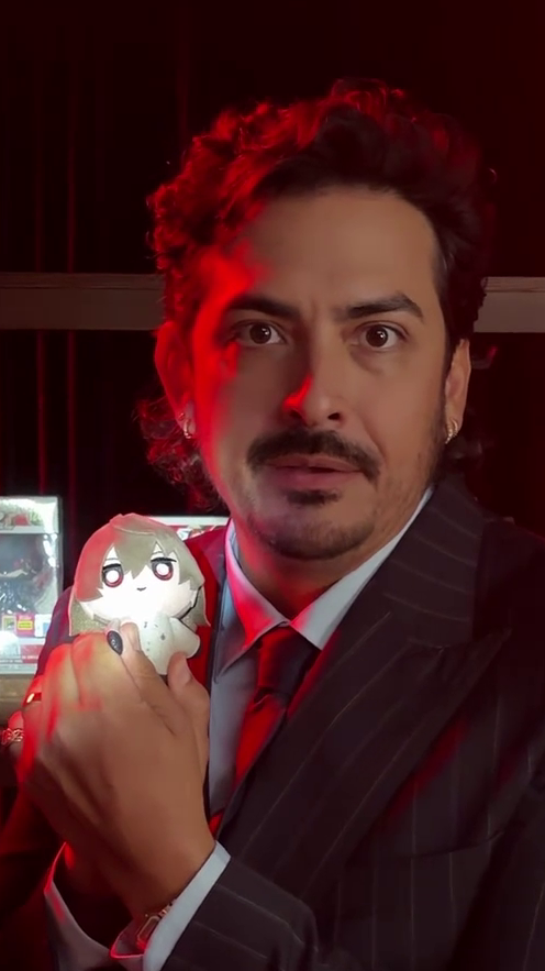 A man with dark curly hair and a mustache, wearing a pinstripe suit with a red tie, holds up a small chibi-style plush doll with gray hair, set against dramatic red lighting and a dark background with monitor screens visible behind him. The behind-the-scenes setting and references to voice actors align with the post's focus on showcasing the cast involved in the Atlus "Your Next Game" campaign.