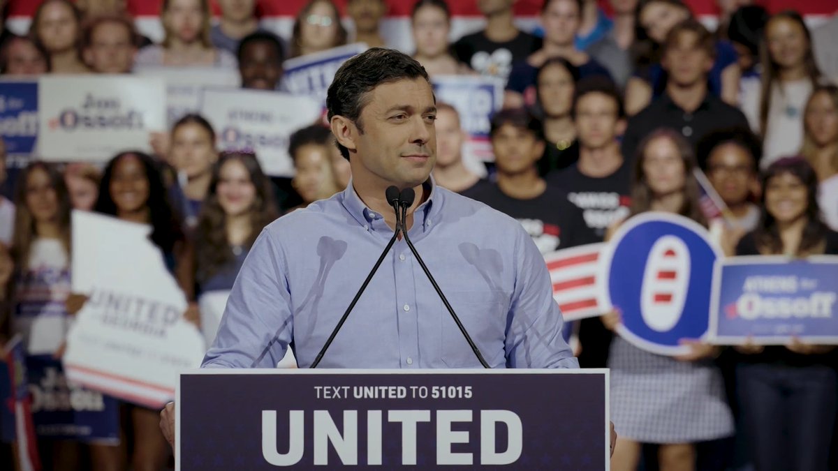 A man in a light blue button-down shirt speaks at a podium labeled "UNITED" with "TEXT UNITED TO 51015," surrounded by supporters holding campaign signs including "Jon Ossoff" and "Ossoff for Senate," consistent with his campaign pitch about reaching out to Georgia mayors, pastors, and farmers—including Republican Mayor Julie Smith of Tifton—to address local needs like hospital dialysis services.
