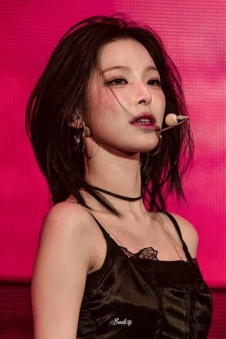 A concert photo of a young woman with short dark hair wearing a black satin top and headset microphone against a bright pink background.