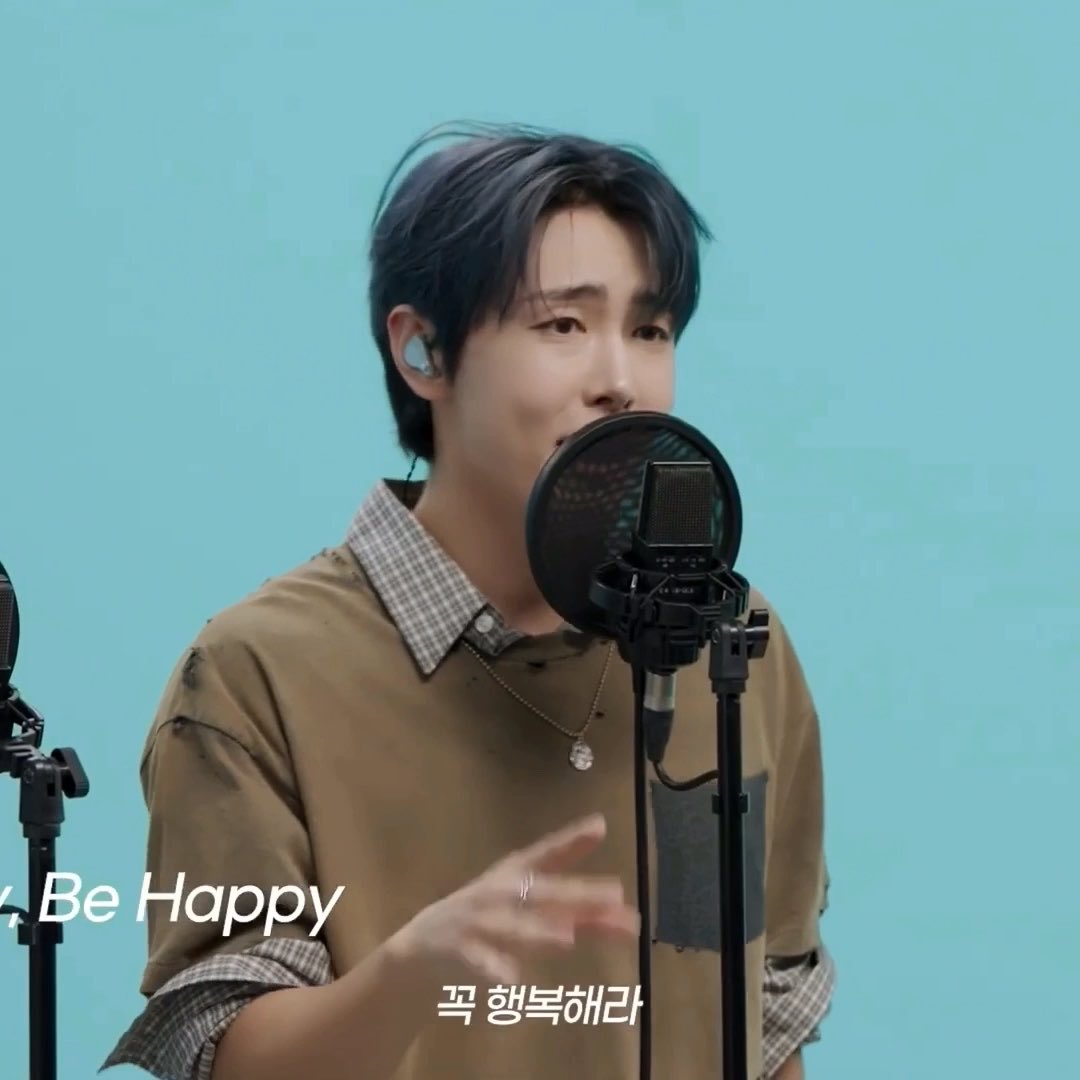 A young man with dark hair sings into a studio microphone wearing a tan shirt over a checkered collar, with the text "Be Happy" and Korean text "꼭 행복해라" visible on screen.