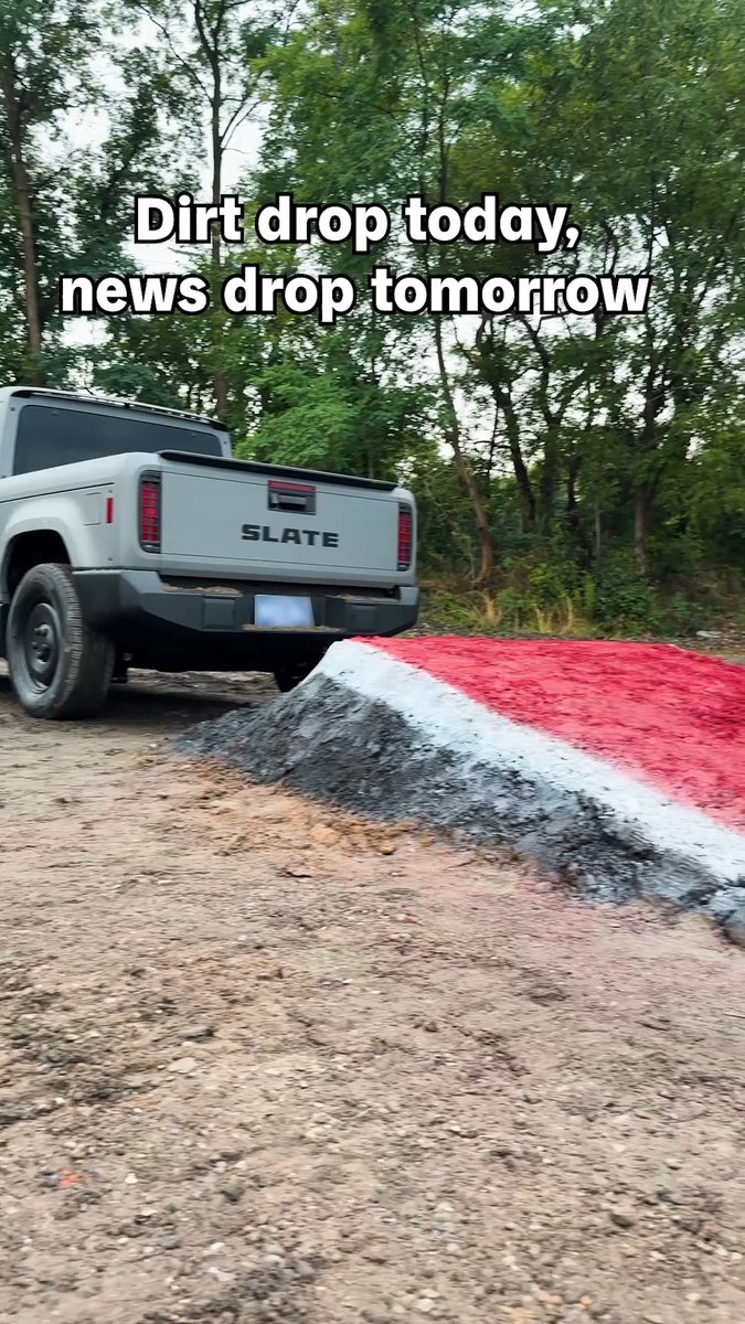 A rear view of a gray Slate electric pickup truck driving off a red, white, and black dirt ramp with text 'Dirt drop today, news drop tomorrow'.