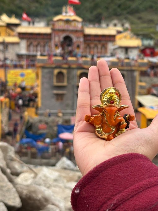 A hand holds a small golden and orange Ganesha figurine against the backdrop of an ornate temple, likely in a mountainous region of India or Nepal.