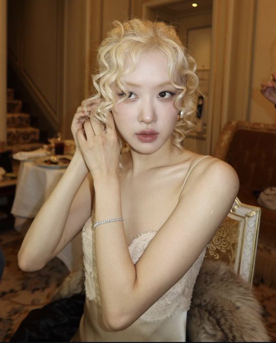 A young woman with blonde curly hair wears an elegant white dress and a diamond bracelet in what appears to be a formal setting.