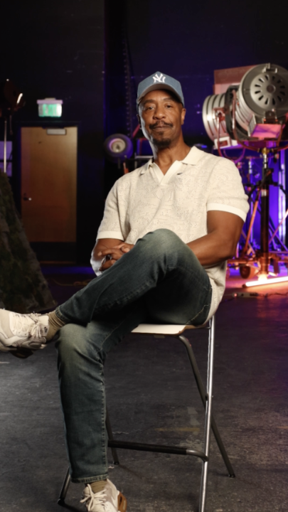 A man wearing a NY baseball cap, white short-sleeved polo shirt, jeans, and white sneakers sits on a stool in what appears to be a studio or theater setting with lighting equipment behind him.