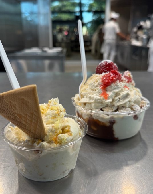 Two cups of ice cream or gelato are displayed on a stainless steel surface in what appears to be a restaurant kitchen, with one topped with raspberries and the other featuring a wafer cookie.
