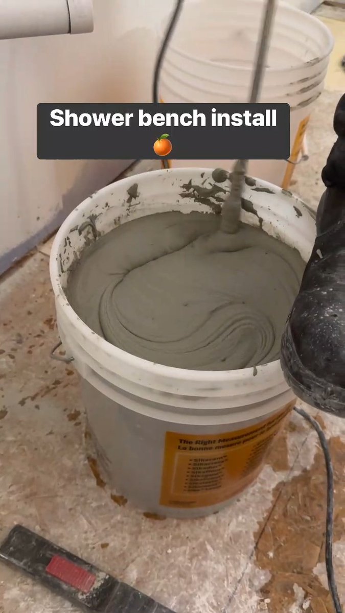 A white bucket containing grey grout being mixed with a drill attachment, overlaid with the text 'Shower bench install 🍊'.