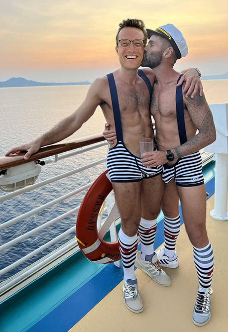 Two shirtless men in matching sailor-themed outfits with striped shorts, suspenders, knee-high socks, and a captain's hat posing together on a ship's deck at sunset, one kissing the other on the cheek.