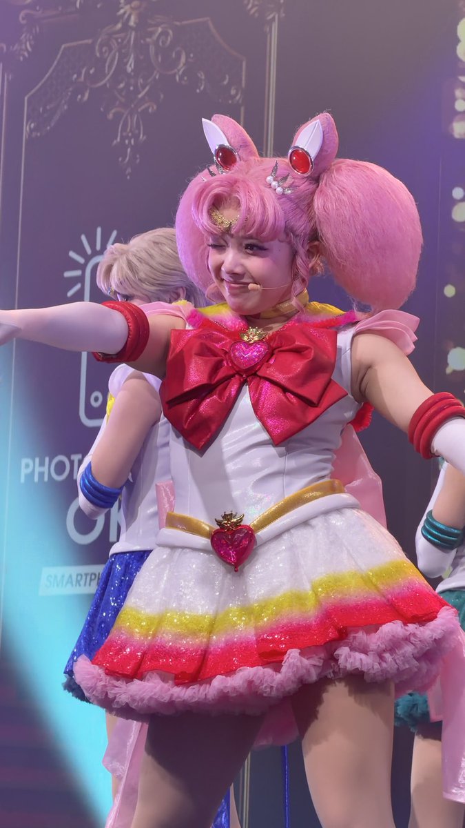 A performer dressed as Chibiusa/Sailor Chibi Moon from Sailor Moon, wearing a pink wig, bunny ear tiara, and a colorful rainbow-frilled sailor outfit on stage.