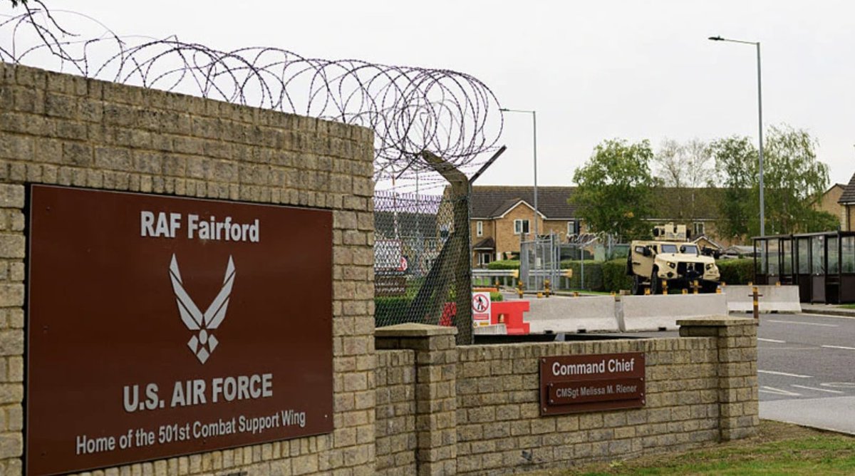 The image shows the entrance to RAF Fairford, a U.S. Air Force base in the UK that serves as the home of the 501st Combat Support Wing, featuring a brick sign, coiled razor wire atop the perimeter wall, and an armored military vehicle stationed at the gate. The accompanying post questions whether a reported foiled terror attack at this British base hosting American troops was a genuine Islamic extremist plot or a staged false flag event intended to draw the UK into broader conflict.