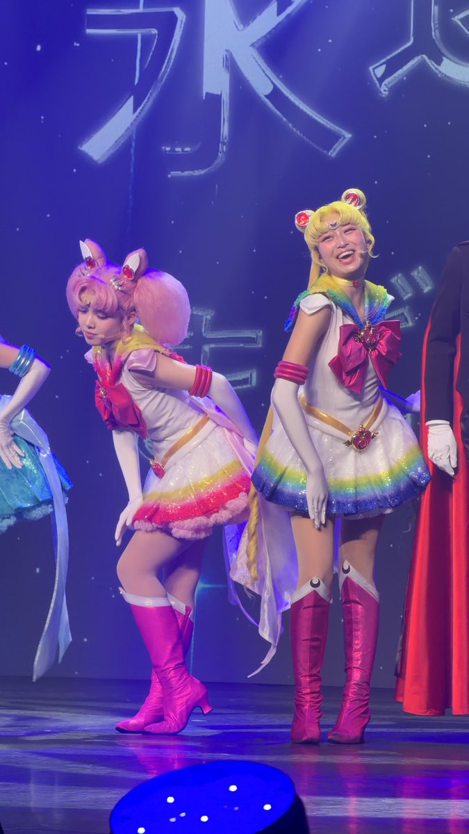 Two performers in Sailor Moon costumes pose on stage, featuring colorful rainbow skirts, pink boots, and character wigs with Japanese characters visible on the backdrop.