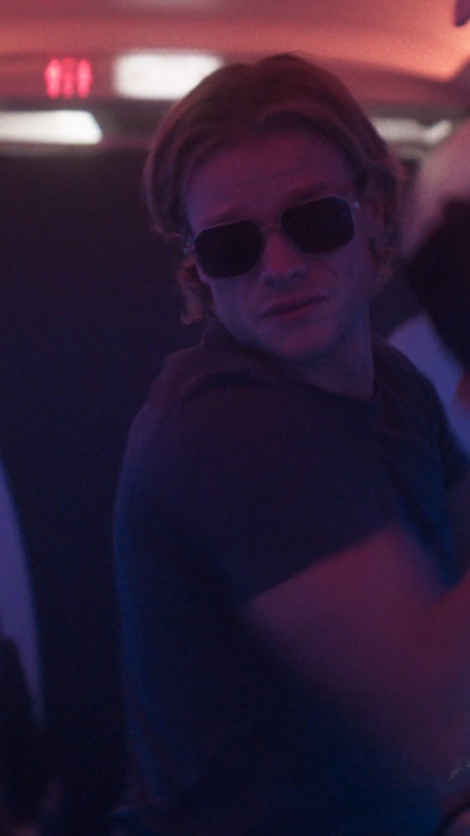 A person with shoulder-length blond hair and dark hexagonal sunglasses glances back over their shoulder in a dimly lit, red-tinged setting that resembles a bar or nightclub scene. The moody, anticipatory atmosphere fits the post's excited countdown to an upcoming date or release.