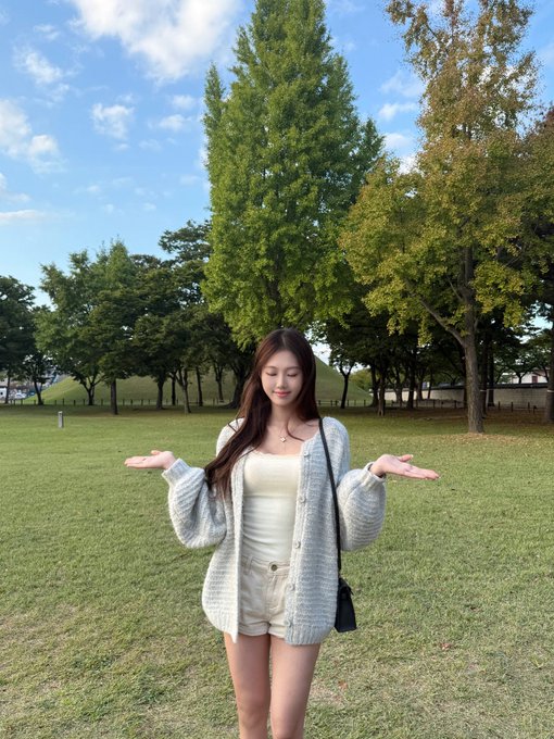 A young woman with long brown hair wearing a cream top and light gray cardigan, standing in a grassy park with trees, holding her hands out.