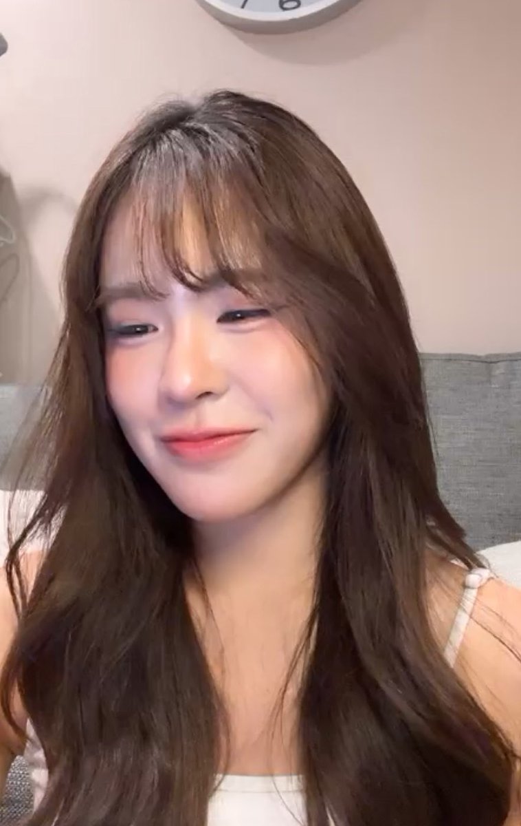 A close-up selfie of a young woman with long brown hair and bangs, smiling gently while wearing a white top, with a clock partially visible on the wall behind her.