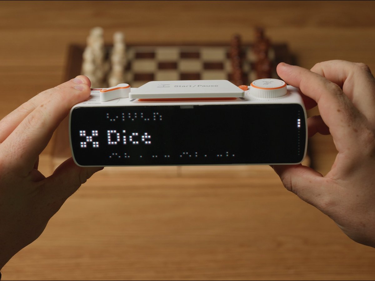 Two hands hold a small white and orange electronic device with a screen displaying pixelated text reading 'X Dice' above what appears to be 'LL LL', set against a wooden table with a chessboard in the background.