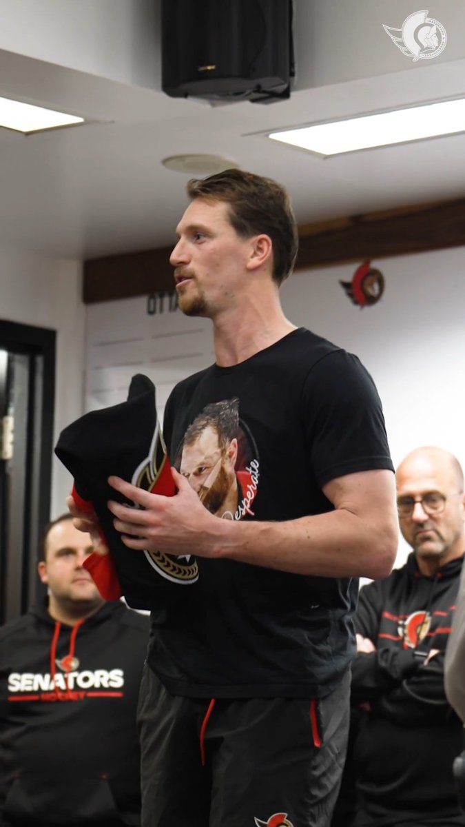 A tall man with a goatee wearing a black t-shirt with a graphic print, standing in a room and holding folded clothing, with other men in 'SENATORS' apparel visible in the background.