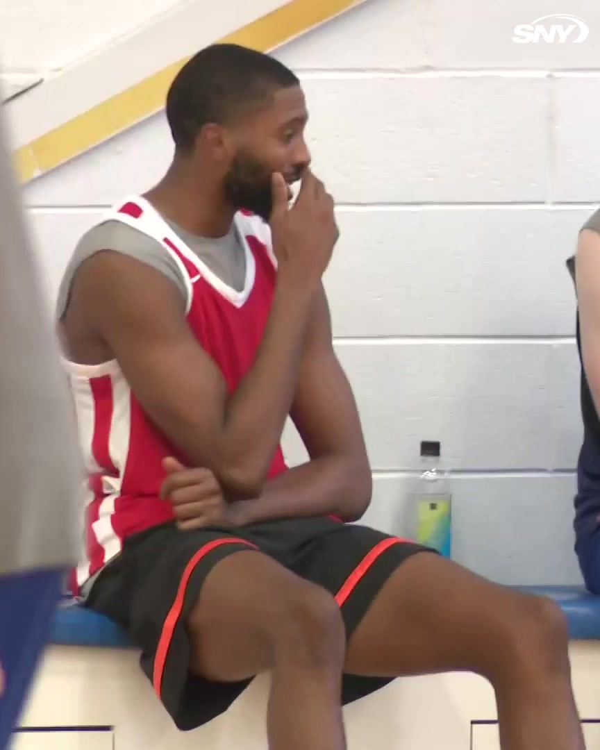 Basketball player Mikal Bridges sits on a bench in a gym wearing a red and white practice jersey, with an SNY logo in the corner.