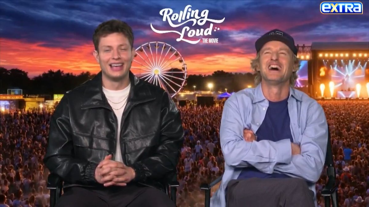 Two men laughing in an interview setting for 'EXTRA' promoting 'Rolling Loud: The Movie,' with a festival background featuring a Ferris wheel, stage, and crowd at sunset.