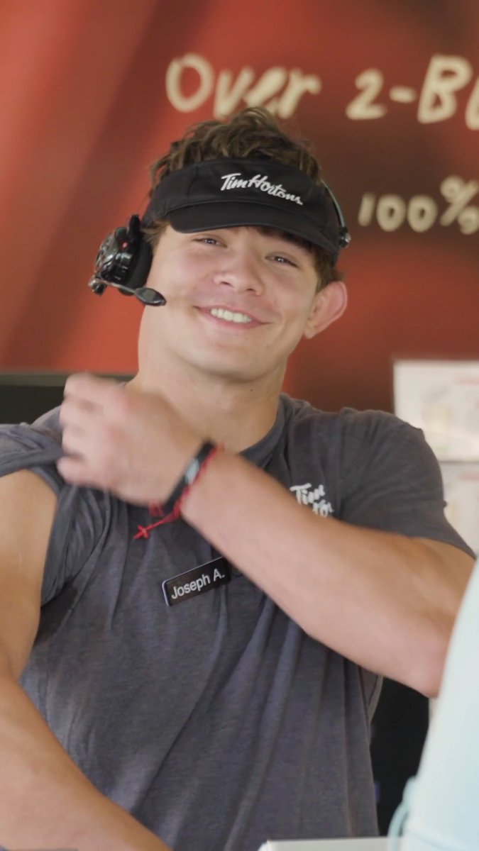 A smiling young Tim Hortons employee wearing a branded visor, headset, and a "Joseph A." name tag stands in front of an "Over 2-B..." promotional sign, fitting the post celebrating National Coffee Day with a playful "Cup of Joe" pun referencing Buffalo Joe / Joe Andreessen.
