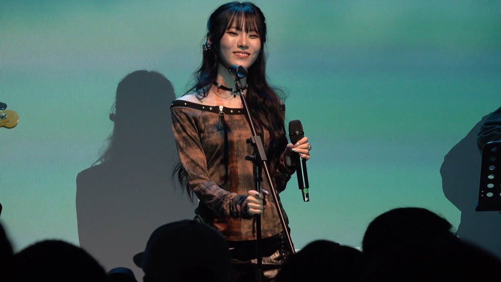 A photograph of a young woman with long dark hair holding a microphone and smiling while performing on stage with a green backdrop.