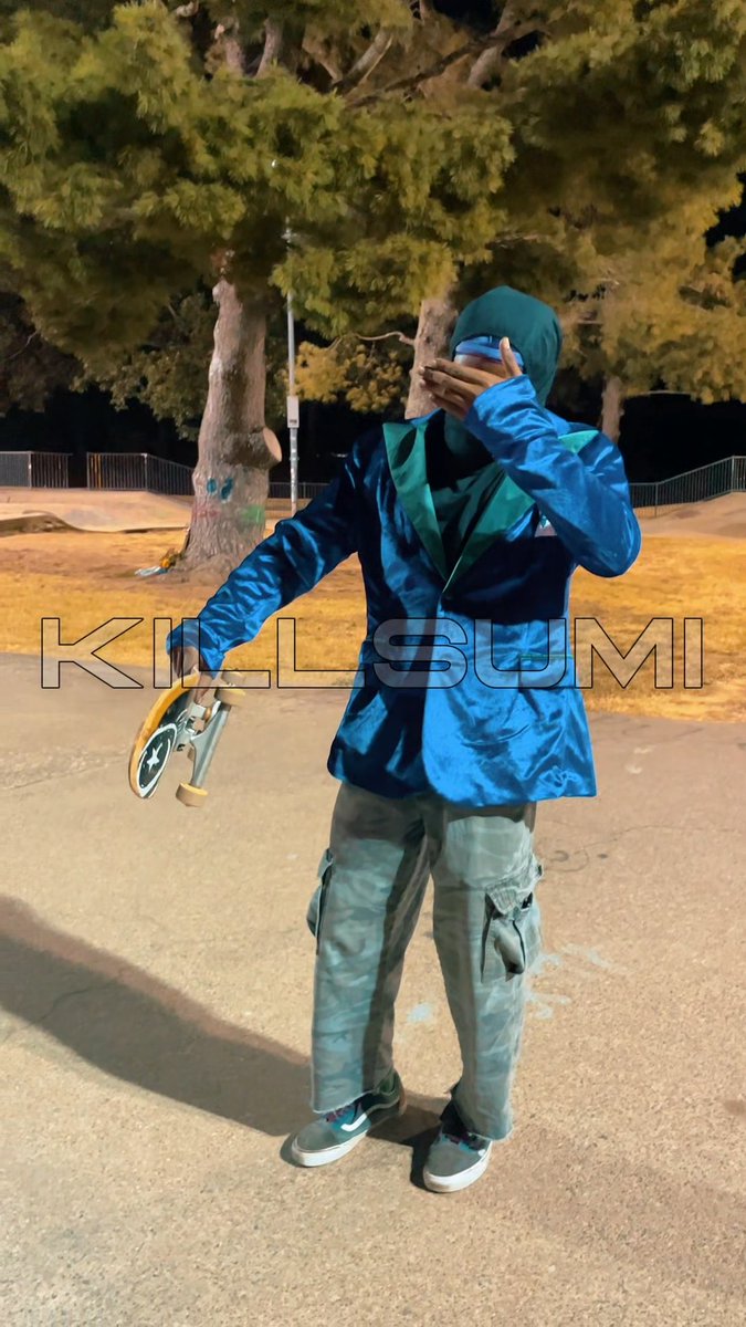 A photograph taken at night shows a person standing in a park wearing a shiny blue jacket with a green star design, gray cargo pants, and a dark hood or mask, holding a small skateboard. The text 'KINLASUMI' is overlaid across the image.