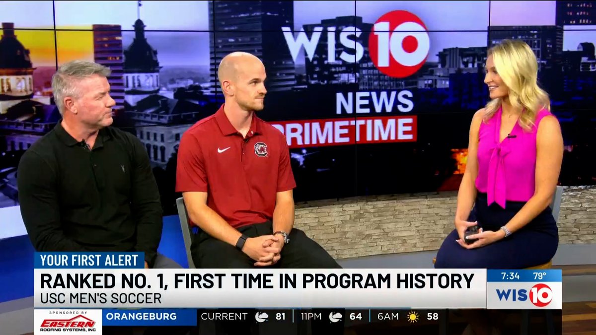 A WIS10 News Primetime broadcast features two men—one in a black shirt and one in a red USC polo—being interviewed by a female anchor, with a chyron reading 'RANKED NO. 1, FIRST TIME IN PROGRAM HISTORY - USC MEN'S SOCCER.'
