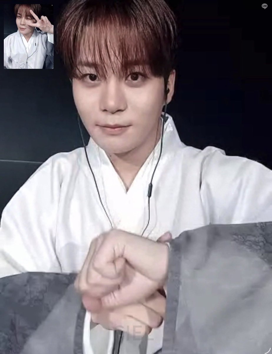 A close-up photo of a young man with brown hair wearing a white traditional Korean hanbok-style top with earphones, taken during what appears to be a livestream with 'CIEL' visible at the bottom.