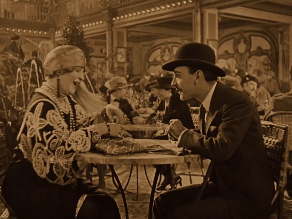 A sepia-toned still from the 1927 silent film *Wings* shows a man in a bowler hat and suit seated across from a woman in an ornate embroidered cap at a small café table, surrounded by other patrons in a bustling Parisian café setting that showcases the famous long tracking shot referenced in the post.