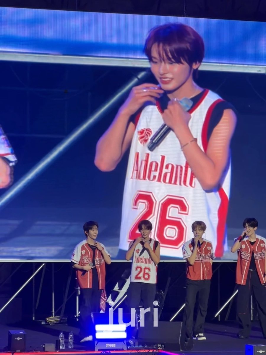 K-pop idol Iuri stands on stage with three groupmates, all wearing matching red and white baseball jerseys; Iuri's jersey number 26 is featured on the large screen behind him.