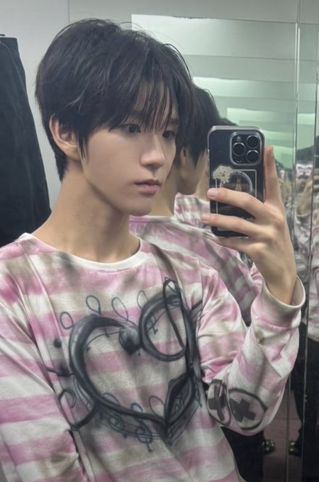 A young man takes a mirror selfie while wearing a pink tie-dye sweatshirt with a heart design.